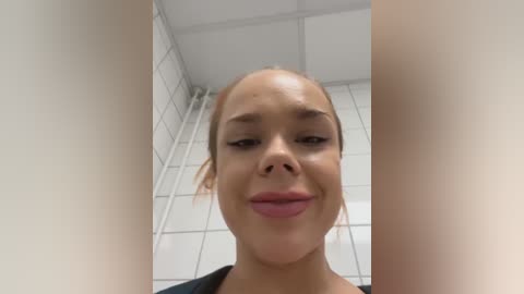 Media: Video of a young woman with light skin and light brown hair, wearing pink lipstick, in a tiled bathroom with white tiles and a white ceiling.