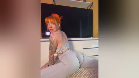 Media: A video of a fair-skinned, red-haired woman with tattoos, wearing tight gray leggings and a sports bra, kneeling on a bed in a dimly lit room with a TV and white dresser.