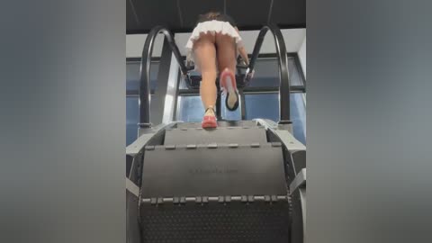 Media: Video of a woman in a short white skirt running down an escalator, showing her bare buttocks. The escalator is industrial with a textured surface and a red emergency stop button.