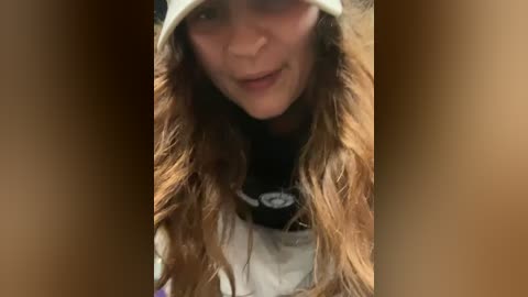 Media: Video of a smiling woman with long, wavy brown hair, wearing a beige bucket hat and a white t-shirt with a black graphic, standing in front of a blurred, wooden background.