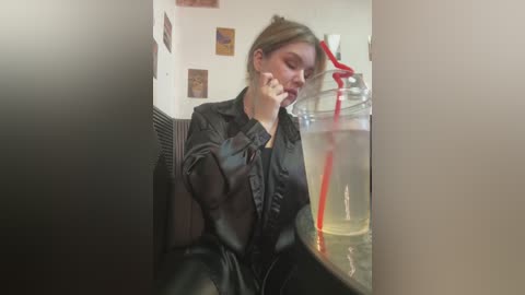 Media: Video of a blonde woman in a black shirt, looking contemplative, seated beside a large glass of milk with a red straw, in a dimly lit cafe with framed artwork on the wall.