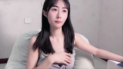 Media: Video of an Asian woman with straight black hair, wearing a sleeveless gray top, sitting on a light gray armchair in a plain, white-walled room.