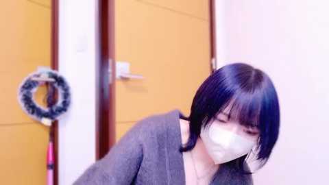 Media: Video of a young woman with short black hair, wearing a gray sweater and a white mask, standing in a beige hallway with wooden doors and a decorative wreath.