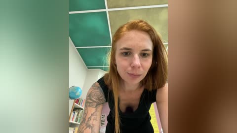 Media: Video of a fair-skinned, red-haired woman with tattoos, wearing a black top, leaning over a table in a brightly lit room with green and beige ceiling tiles.