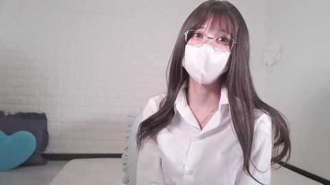 Media: Video of an Asian woman with long dark hair, wearing white glasses and a white mask, sitting indoors with white brick wall and blue plush toy in the background.