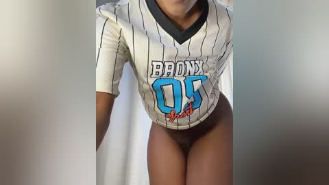 Media: Video of a dark-skinned woman wearing a white and black pinstriped jersey with \"BROOKLYN 03\" on it, revealing her black pubic hair. The background is blurry, with neutral tones.