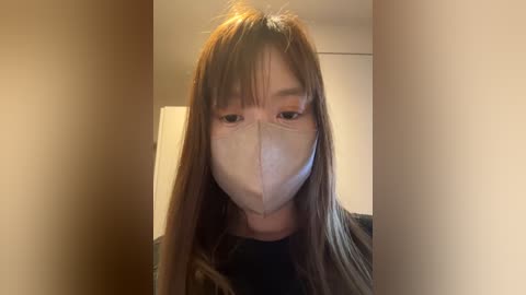 Media: Video of a young woman with long brown hair, wearing a beige face mask, black top, and a white door in the background.