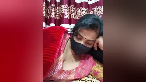 Media: A video of an Indian woman in a red sari with a black face mask, lying on a bed with a floral-patterned sheet. She has a slender build and medium-length black hair, resting her head on her hand.