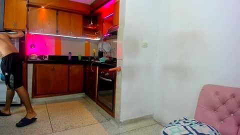 Media: Video of a modern kitchen with wooden cabinets, black countertops, and pinkish ambient lighting. Person in blue shorts and sandals stands near the sink, facing away from the camera.