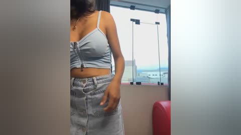 Media: Video of a woman in a light blue, off-the-shoulder crop top and high-waisted light blue denim shorts, standing indoors by a window with a red chair and airport view in the background.