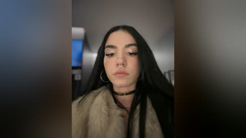 Media: Video of a young woman with long black hair, pale skin, and dark eyebrows, wearing a fur coat, choker, and hoop earrings, set against a blurry background.