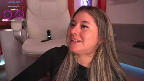 Media: Video of a smiling, light-skinned woman with long, straight blonde hair, wearing a black top, sitting in a modern bathroom with a white toilet and pink curtains in the background.