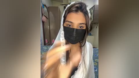 Media: Video of a young woman with medium brown skin, brown hair, and a black face mask, holding a silver hairbrush. She is indoors, wearing a white robe, in a room with a floral carpet and a wooden dresser.