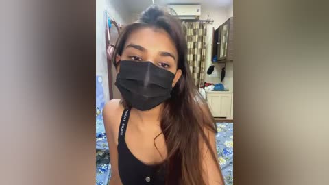 Media: A video of a young woman with medium skin tone, long brown hair, and a black mask, wearing a black tank top, standing in a cluttered kitchen with beige walls and floral curtains.