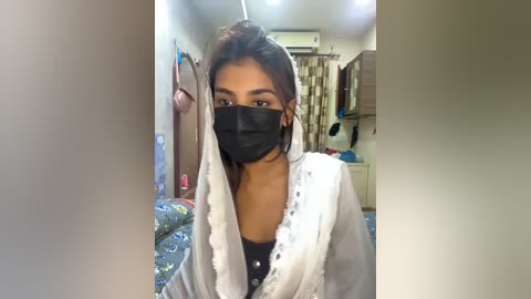 Media: Video of a young South Asian woman with medium brown skin, wearing a black face mask, white shawl, and black top, in a cluttered, dimly lit bedroom with floral-patterned bedspread, clothes hanging, and a window with curtains.