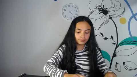 Media: Video of a young girl with long black hair, wearing a black-and-white striped shirt, sitting in front of a white wall with black floral and clock decals.