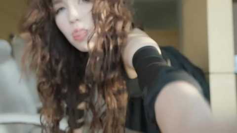 Media: A blurry video of a young Asian woman with long, curly brown hair, wearing a black off-the-shoulder top, sticking her tongue out playfully. Background features a beige wall and a white chair.