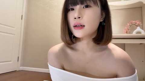 Media: Video of an Asian woman with straight, shoulder-length brown hair and fair skin, wearing a white off-shoulder top. She sits in a beige room with a closed white door, wooden floor, and a white vanity with pink flowers.