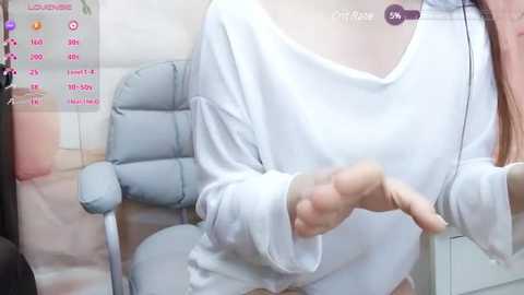 Media: Video of a woman wearing a loose, off-shoulder white sweater, sitting on a light grey chair in a modern room with a pink plant and a white wall.