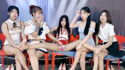 Media: Video of five Asian women in revealing, colorful, traditional and casual attire, sitting on red plastic chairs in a studio setting.