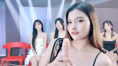 Media: Video of an Asian woman with long, straight hair, wearing a black dress, holding a large, erect penis in front of a modern, minimalist room with white walls and red chairs.