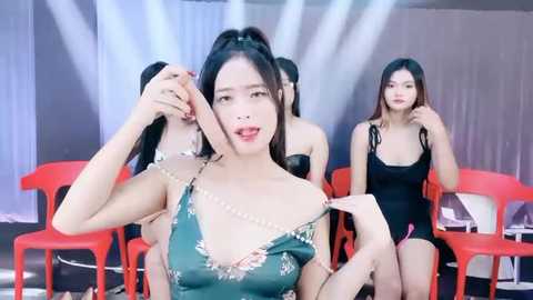 Media: Video of four Asian women in a studio setting. Central woman, with long black hair, wearing a revealing green dress, poses seductively. Background includes red chairs, white curtains, and studio lighting.