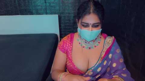 Media: Video of a South Asian woman in a colorful sari with a low neckline, wearing a face mask, sitting on a dark bed against a textured green wall.