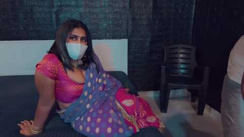 Media: Video of a woman in traditional Indian attire, wearing a pink and blue saree with gold embroidery, seated on a bed with a black headboard, masked, and handcuffed. Background features dark curtains and a black plastic chair.