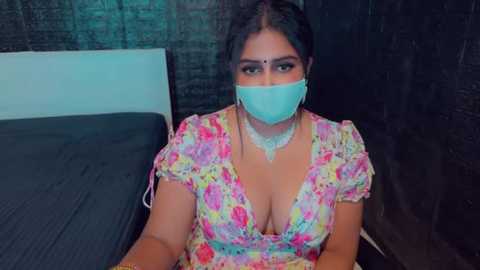 Media: Video of a South Asian woman with dark hair, wearing a floral dress and a face mask, seated on a bed in a dimly lit room with dark brick walls.