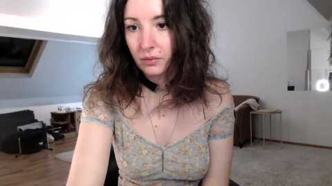 Media: Video of a woman with curly brown hair, wearing a floral off-shoulder top, in a modern, minimalist living room with light wooden floors and white walls.