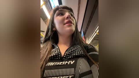 Media: Video of a young woman with long brown hair, wearing a black and white patterned shirt, taken from a low angle in a subway car.