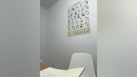 Media: Video of a minimalist, white-walled room with a white chair, a wooden desk, and a poster of the alphabet and numbers on the wall.