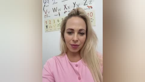 Media: Video of a blonde woman with fair skin, wearing a pink shirt, standing in front of a classroom wall adorned with alphabet and number charts. She has a subtle smile.