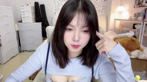 Media: A video of an East Asian woman with fair skin, shoulder-length black hair, and a light blue top. She's in a cozy bedroom with white furniture and a stuffed bear.