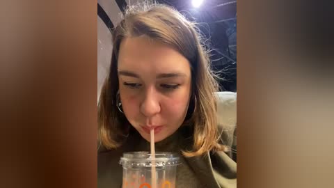 Media: A candid video of a young girl with light brown hair, wearing headphones, sipping from a glass jar filled with orange liquid, in a dimly lit indoor setting with black and white stripes in the background.