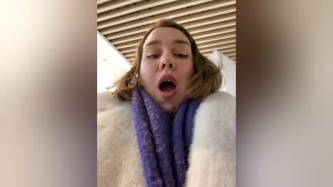 Media: A video captures a young Caucasian woman with light brown hair and a flushed face, wearing a white and purple towel, leaning against a beige wall with horizontal wooden slats.