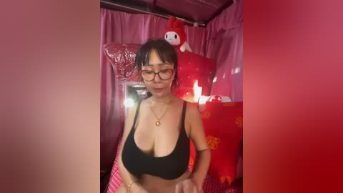 Media: A video of an Asian woman with short black hair, glasses, and a black tank top, standing in a room with red and pink decor, including a large red inflatable character.