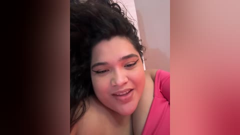 Media: Video of a plus-size Latina woman with curly black hair, wearing a pink top, smiling softly while listening to white earbuds.