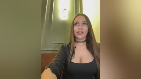 Media: Video of a fair-skinned woman with long, straight brown hair, wearing a black top and choker, sitting indoors with green walls and a wooden chair.