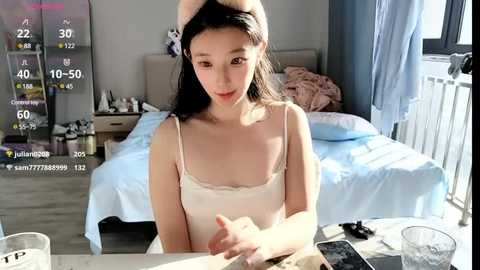 Media: Video of a young Asian woman with long black hair, wearing a white lace-trimmed camisole, sitting at a table in a modern bedroom with a neatly made bed, a white nightstand, and a window.