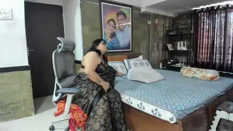 Media: Video of a woman in a black dress sitting on a bed with patterned sheets, framed picture of a family on the wall, and a chair with a red and orange blanket.