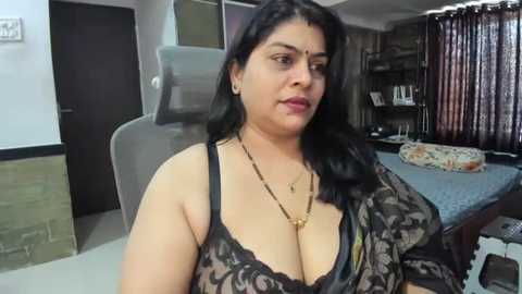 Media: Video of a South Asian woman with long black hair and medium complexion, wearing a black lace bra, sitting in a living room with a beige couch, wooden furniture, and patterned curtains in the background.