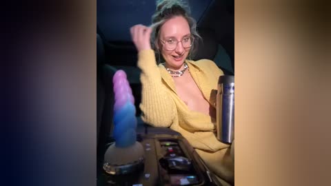 Media: A video of a smiling, fair-skinned woman with glasses and a yellow cardigan, sitting in a dimly-lit car. She has a messy bun and is holding a pink and blue dildo, surrounded by other sex toys on the car's center console.