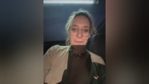 Media: Video of a woman with fair skin, glasses, and brown hair in a car, wearing a beige jacket over a black top. Background shows dim, dark car interior.