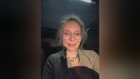 Media: Video of a smiling woman with glasses, messy hair, and a beige jacket, taken inside a car with dim lighting.