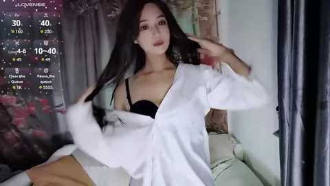 Media: Video of an Asian woman with long black hair, wearing a white shirt and black bra, seated on a white couch, with a digital clock and weather display in the background.