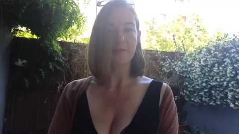 Media: Video of a Caucasian woman with shoulder-length, straight brown hair, wearing a black top and a brown cardigan, standing outdoors against a sunlit garden with greenery and a wooden fence in the background.