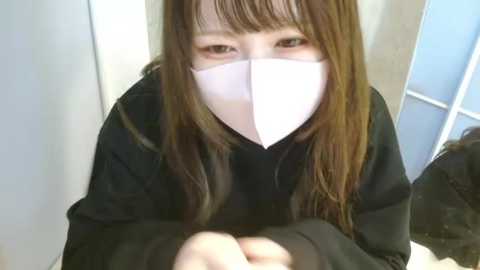 Media: A video of an East Asian woman with long, straight brown hair, wearing a white surgical mask and a black hoodie, kneeling in a white-walled room with a blue door.