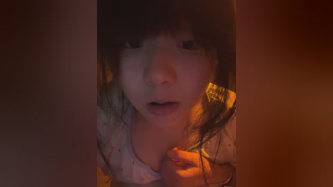 Media: Video of a young Asian girl with dark hair and bangs, wearing a white shirt, in a dimly lit room, eyes wide, mouth open in surprise.