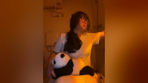 Media: Video of a young woman with long dark hair, wearing glasses, a white t-shirt, and holding a black and white panda plushie. She's in a dimly lit room with a medical device in the background.